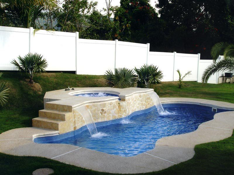 A backyard pool with a waterfall, hot tub, and stone accents, surrounded by green grass and a white fence.