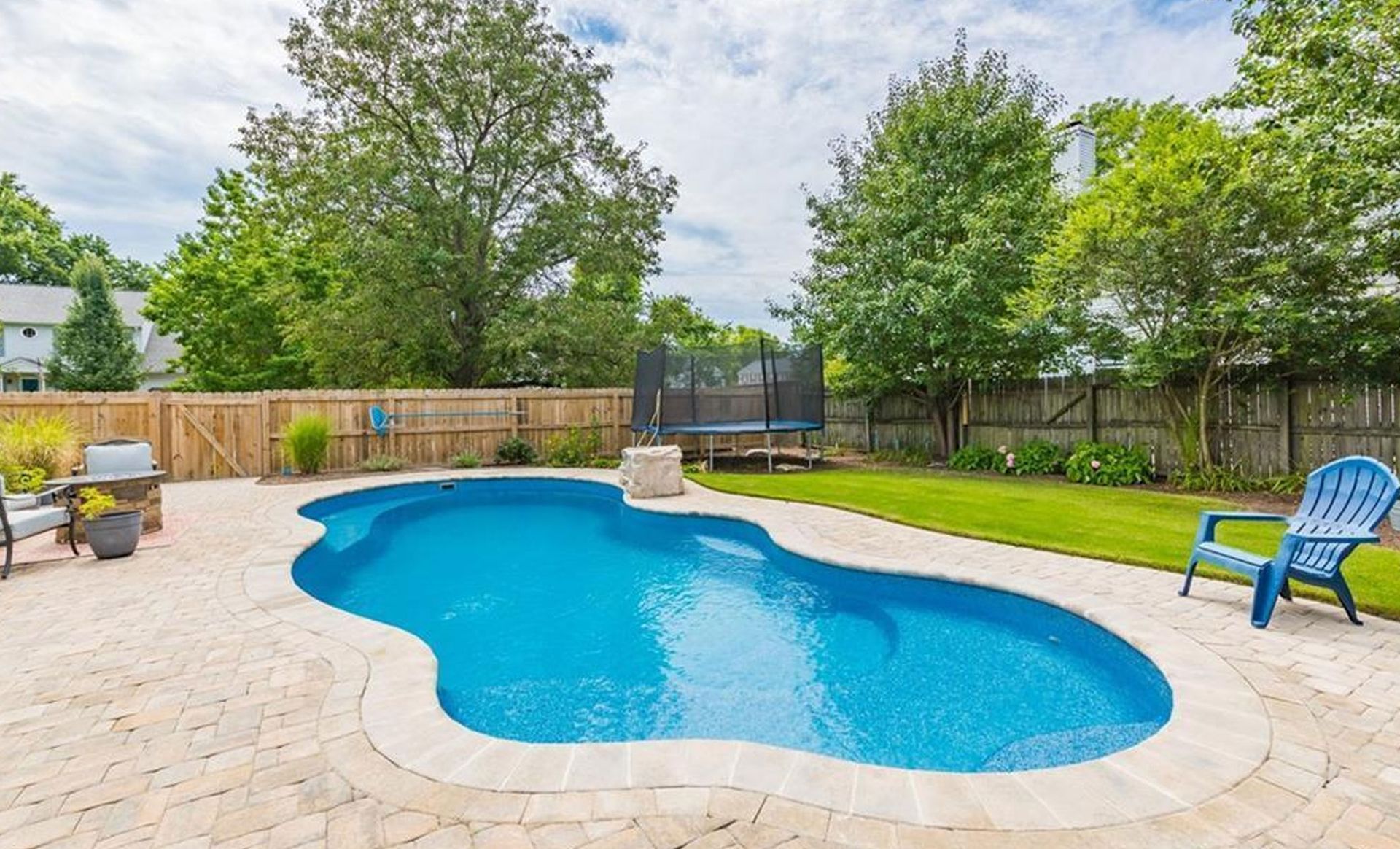 Backyard with a blue swimming pool, brick patio, green grass, and wooden fence.
