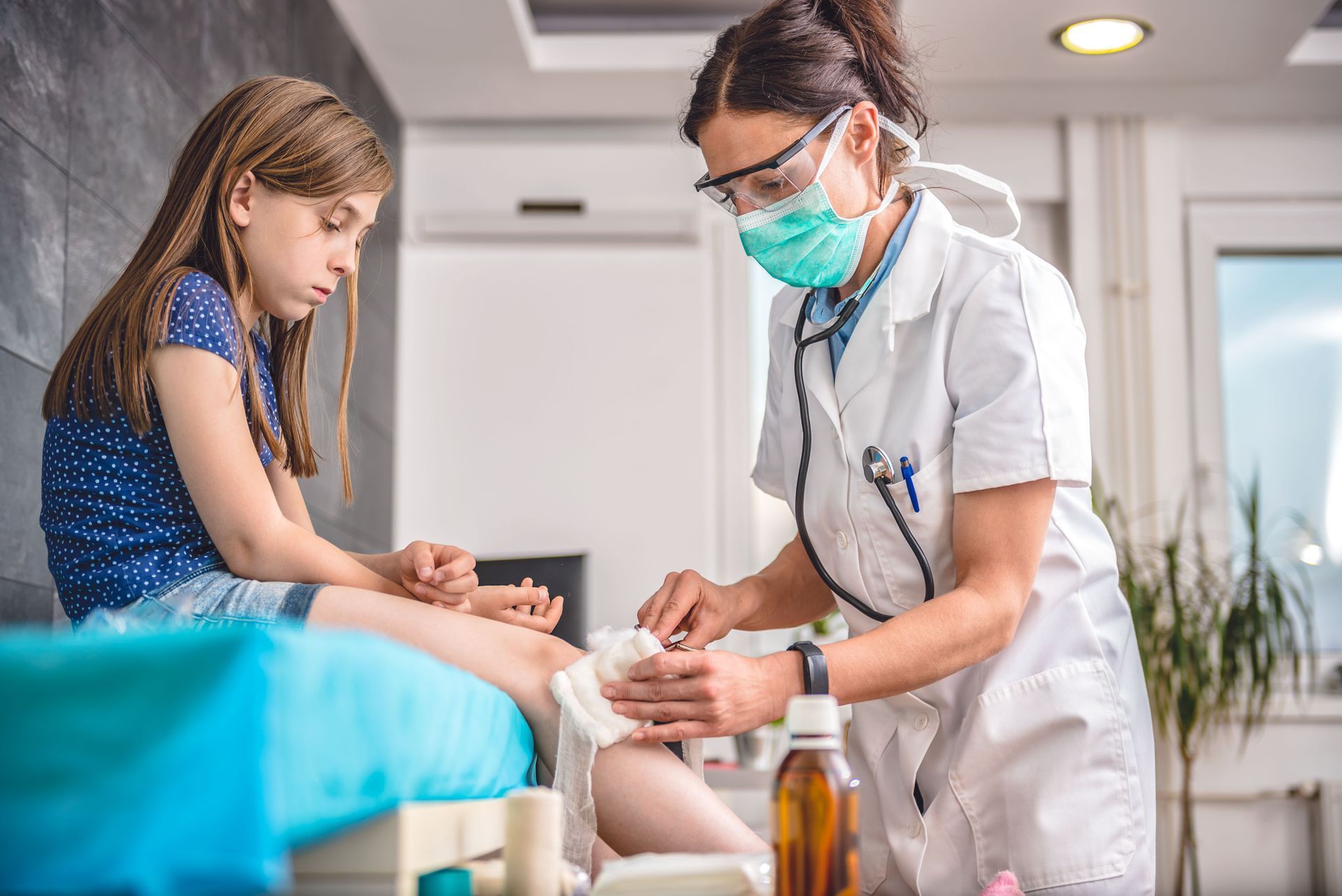 Doctor bandaging a girl's leg wound in a medical office; doctor wears mask and glasses, girl looks concerned.