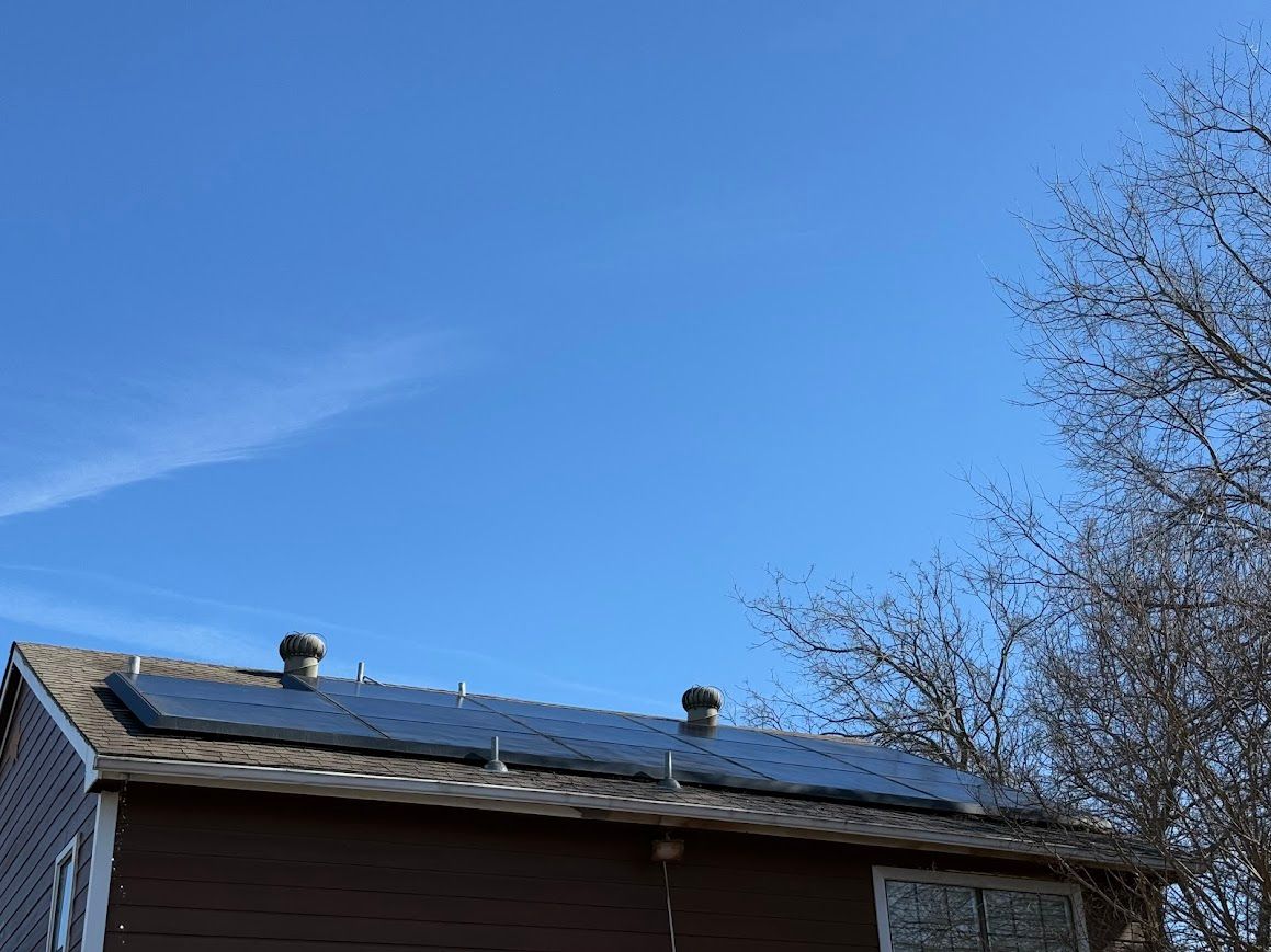 Solar panels on a brown house roof against a bright blue sky with wispy clouds and a tree.