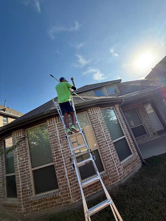 Man on ladder pressure washing a brick house roof under a sunny sky.