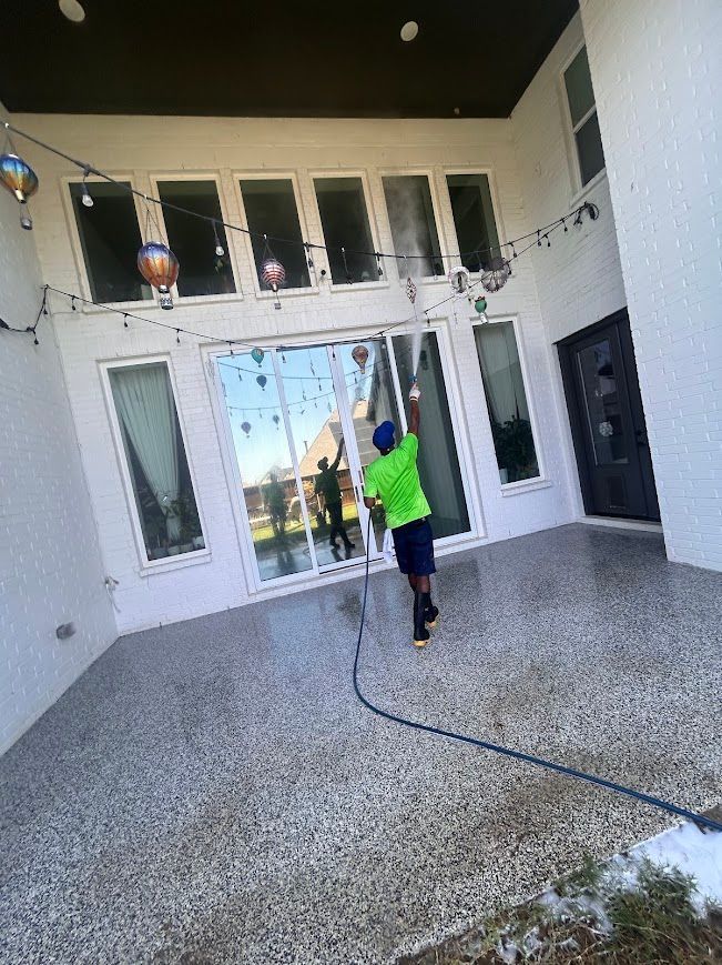 Person power washing a patio with a hose, in front of a white building with windows.