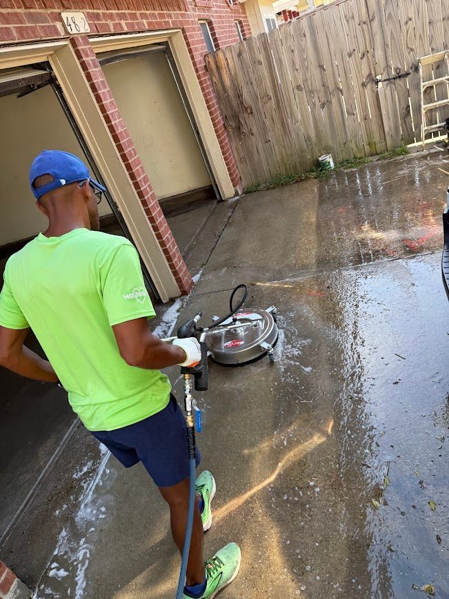 Person in green shirt pressure washing a driveway near a garage, light-colored concrete.