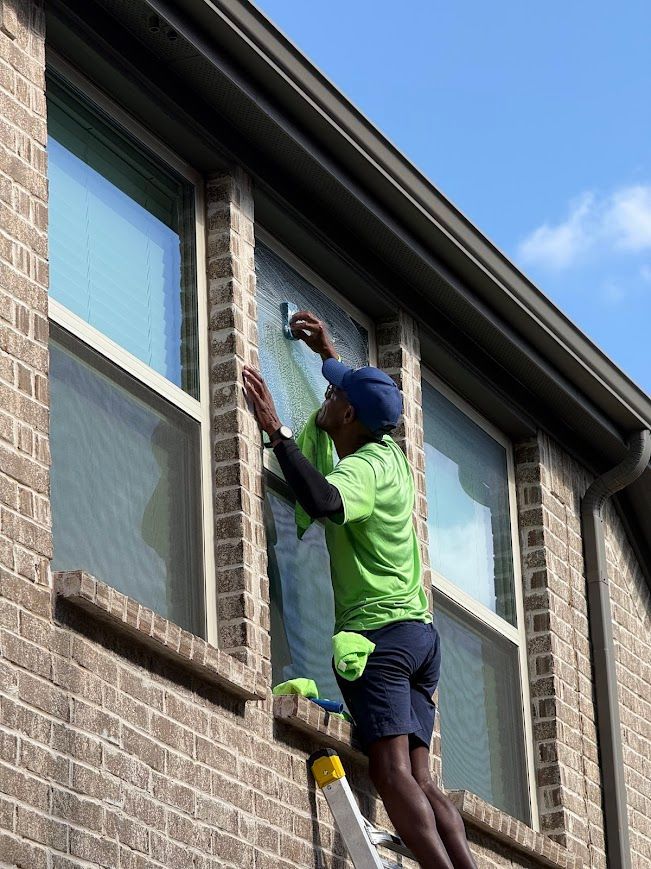 Person on ladder applying film to a window, outdoors. The person wears green shirt, blue hat, and is near a brick building.