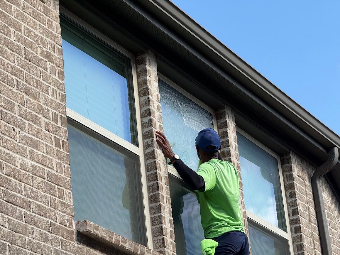 Person washing a window on a brick building, wearing a green shirt and blue cap.