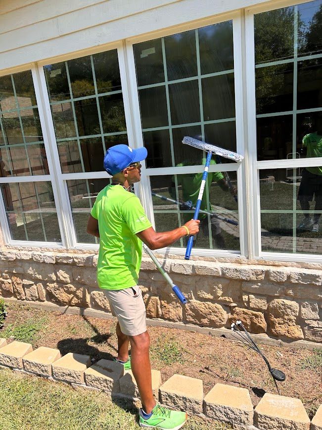 Person cleaning a large window with a squeegee. Wearing a green shirt, blue hat, and khaki shorts. Outdoors.