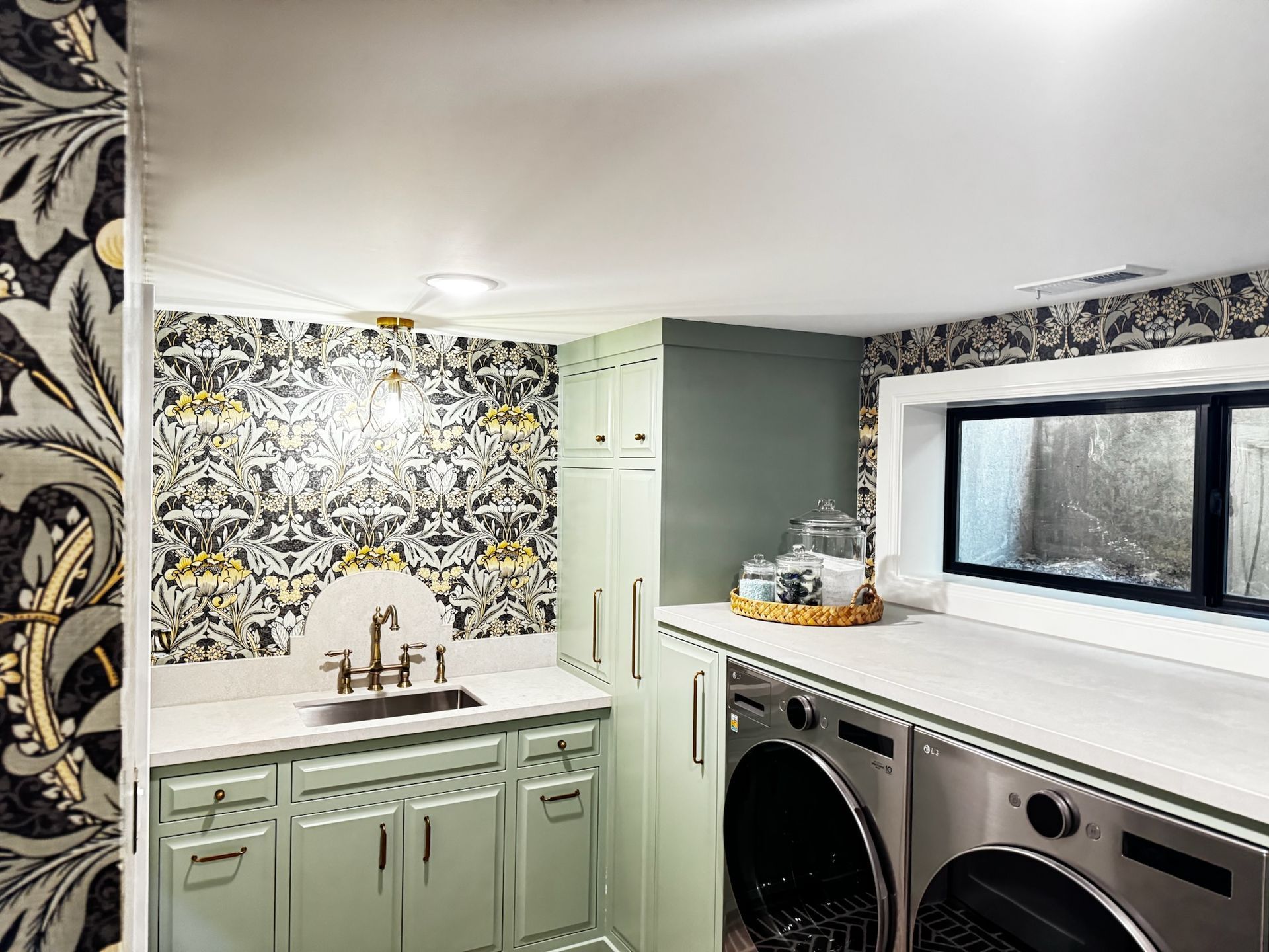Bright laundry room with mint cabinets, patterned wallpaper, sink, countertop, and stacked washer and dryer