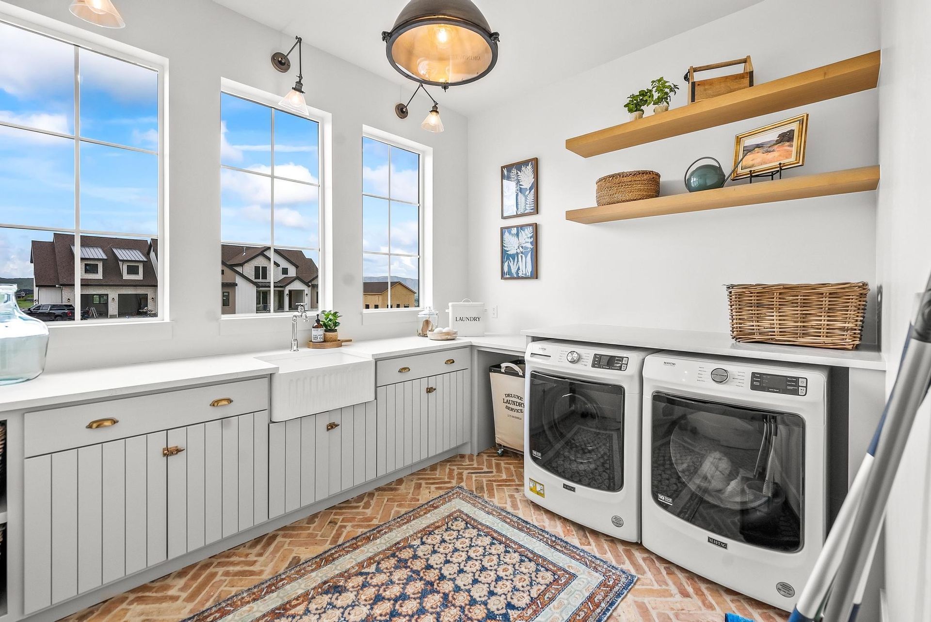Bright laundry room with front-load washer and dryer, white cabinets, wood shelves, and patterned rug.