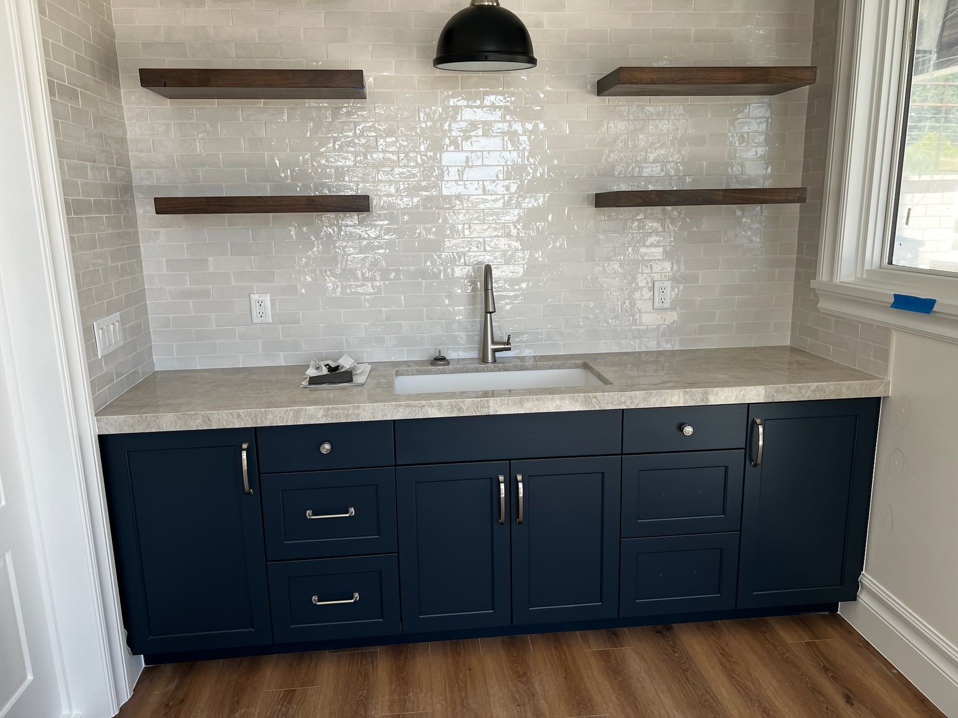 Modern kitchen wet bar with navy cabinets, white countertop, wall shelves, sink, and black pendant light