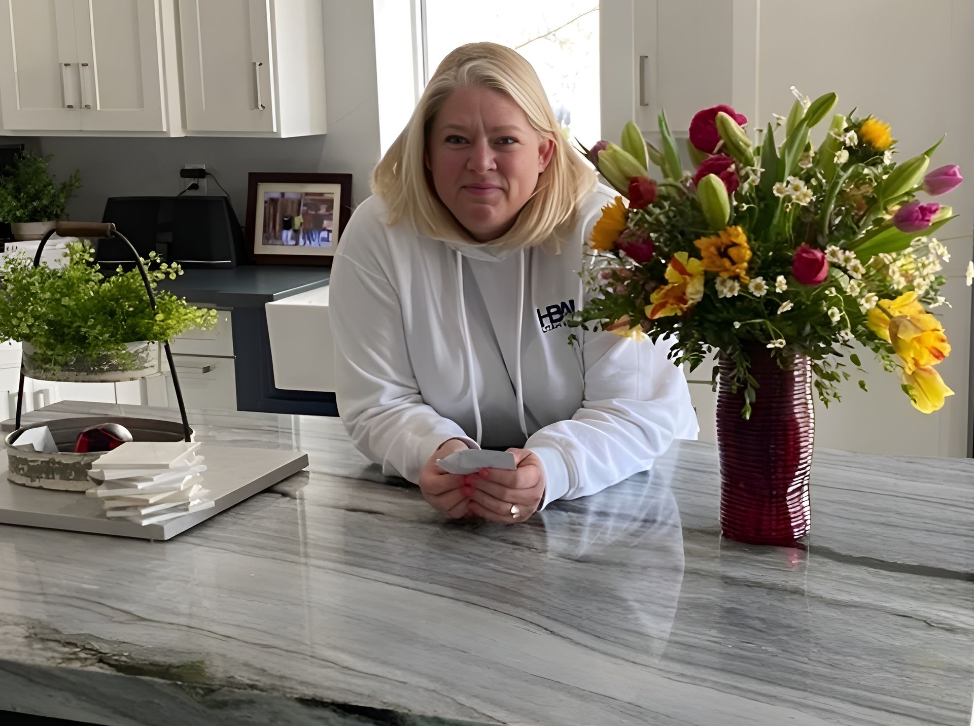 Woman in white hoodie leans on granite countertop in a white kitchen with a flower arrangement.