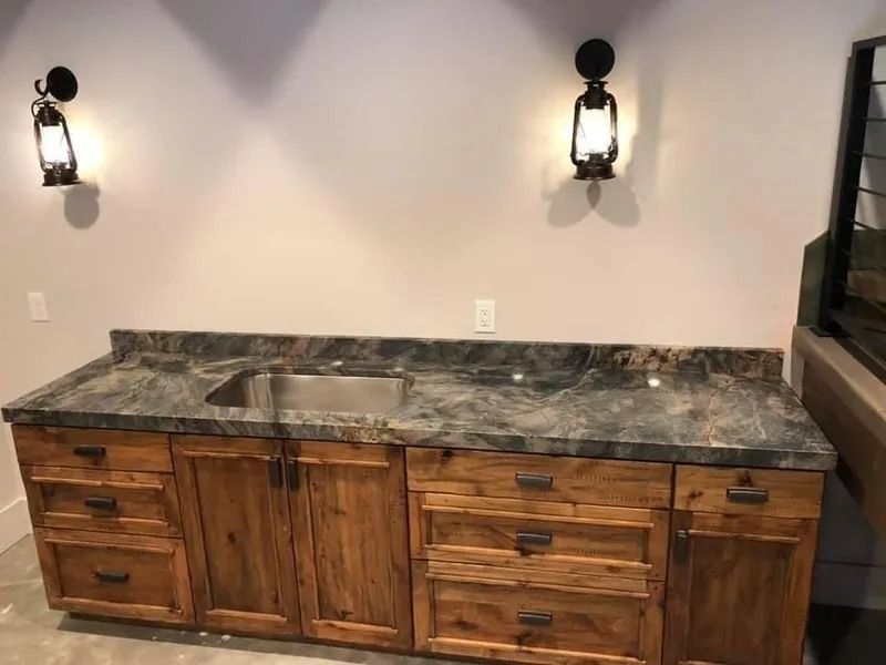Rustic wooden bar with gray countertop, stainless steel sink, cabinets, drawers, and two lantern sconces.