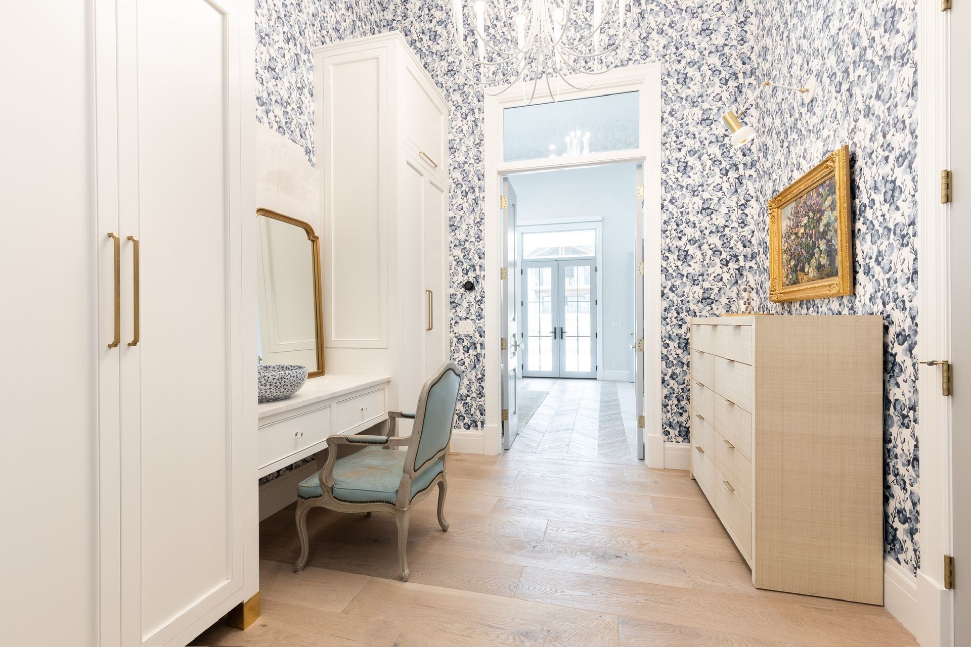 Elegant hallway with blue floral wallpaper, white wardrobes, a vanity chair, and a dresser with a gold-framed mirror
