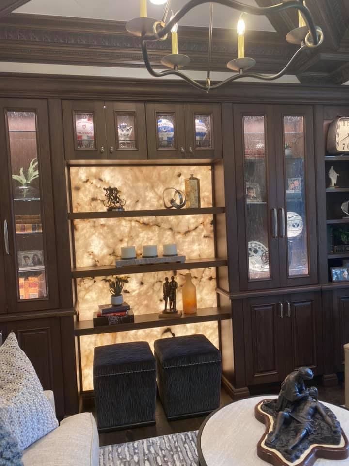 Dark wood built-in bar cabinet with glass doors, backlit shelves, stools, and a chandelier overhead