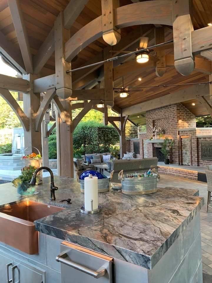 Rustic outdoor kitchen with stone island, copper sink, wood beams, and ceiling fans under a covered patio