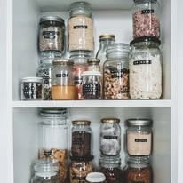 A white cupboard with two shelves filled with labeled glass storage jars containing various pantry ingredients.