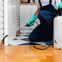 A pest control technician in dark blue overalls and green gloves sprays liquid along a doorway with a portable tank.
