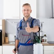 A pest control technician in a grey uniform and plaid shirt gives a thumbs up while holding a chemical sprayer in a kitchen.
