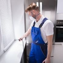 A person wearing a white shirt, blue overalls, and a face mask uses a black cable tool near a window in a kitchen.