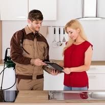 A service technician in brown workwear shows a tablet to a woman in a red shirt inside a modern kitchen.