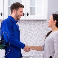A pest control technician in a blue uniform shakes hands with a person in a kitchen with white cabinets and tiled walls.