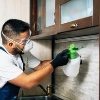 A person wearing a face mask and protective gloves sprays a cleaning solution onto a kitchen backsplash.
