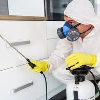 A pest control technician in a white protective suit, mask, and yellow gloves sprays chemicals near white kitchen cabinets.