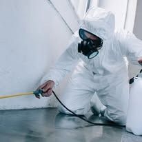 A person in a white protective hazmat suit and a respirator mask sprays a cleaning solution against a wall.