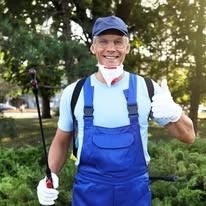 A smiling pest control worker in blue coveralls, gloves, and a face mask holds a sprayer and gives a thumbs up outdoors.