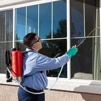 A person wearing a backpack sprayer and green gloves sprays a solution onto a residential window.