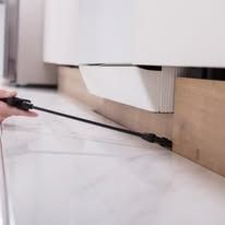 A hand uses a long, black spray wand to apply pesticide along the base of white kitchen cabinets.