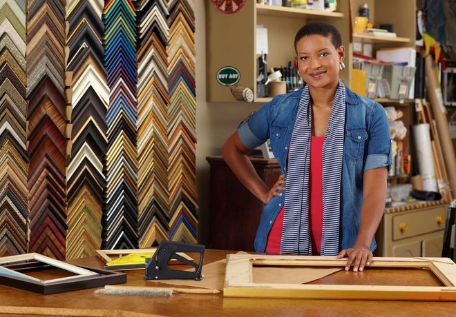 Person standing at a workshop table with fabric swatches and framed wood pieces in the background
