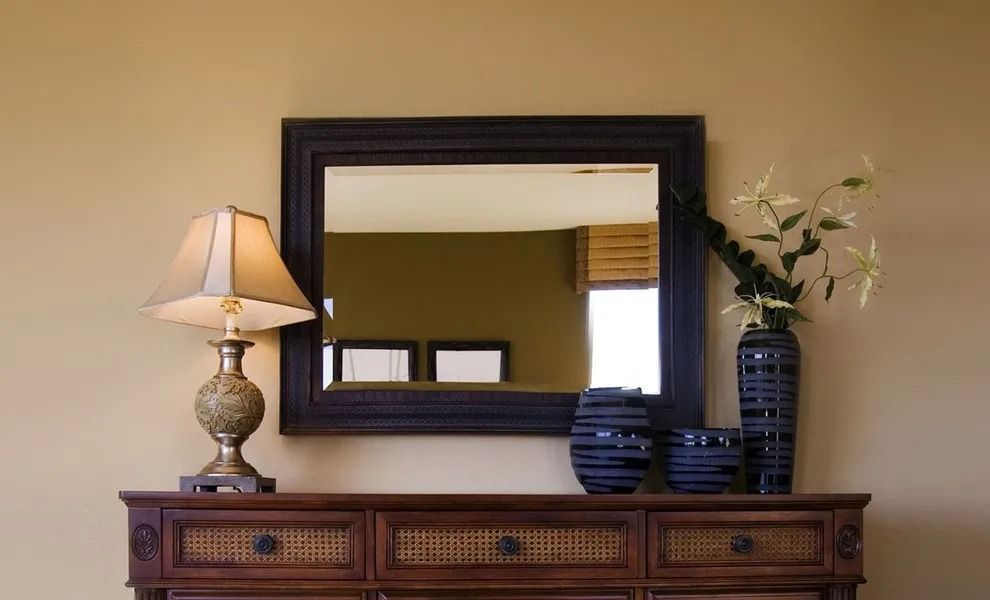 Wooden dresser with lamp, black mirror, stacked plates, and white flowers against a beige wall