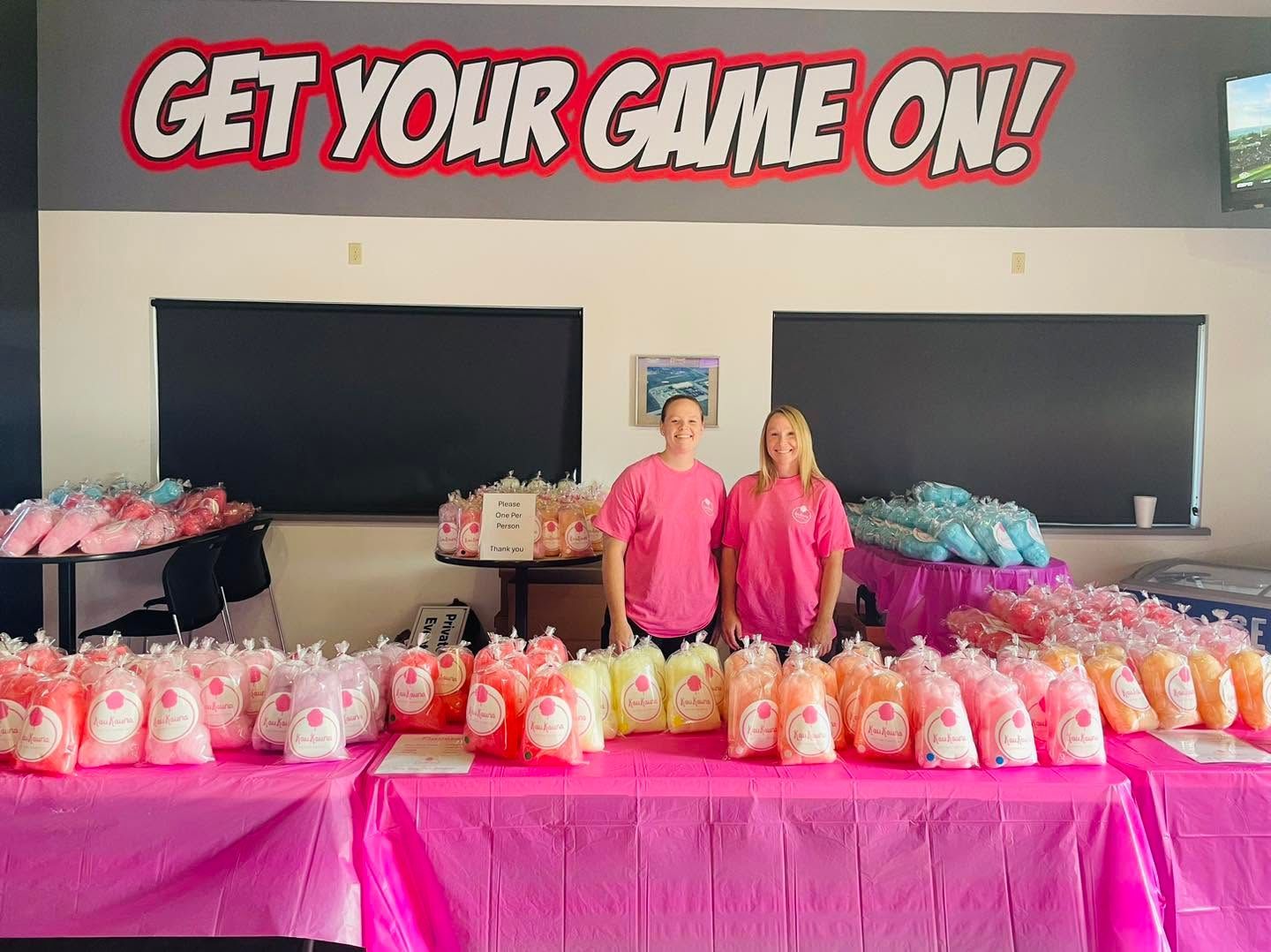Two people behind a pink snack table under a “GET YOUR GAME ON!” banner