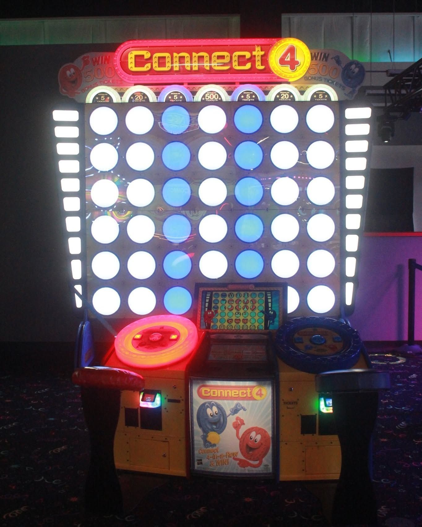 Connect 4 arcade game with large lit grid and colored control stations in an indoor arcade