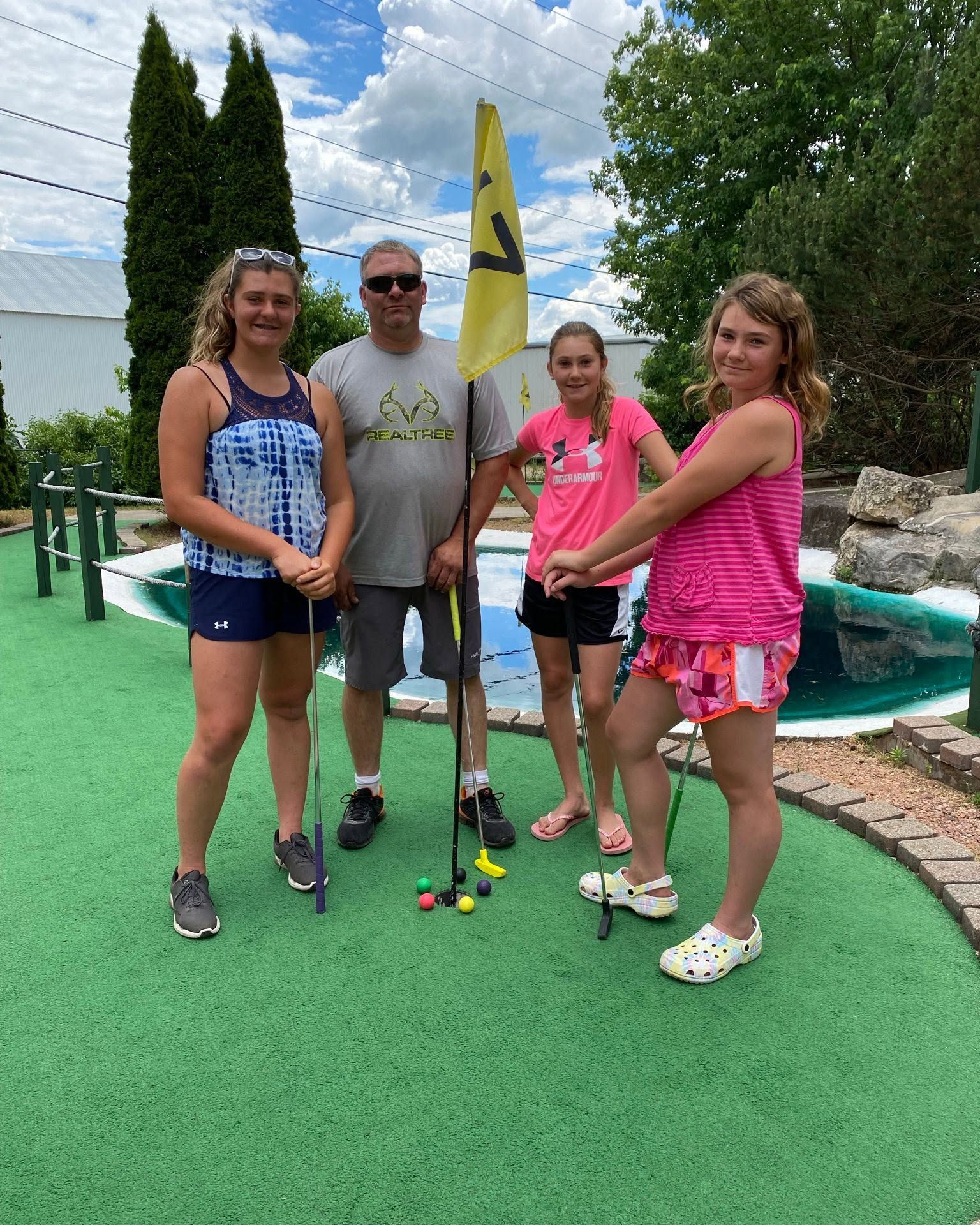 Four people posing with mini golf clubs on a green putting course, with a yellow flag and water feature behind them