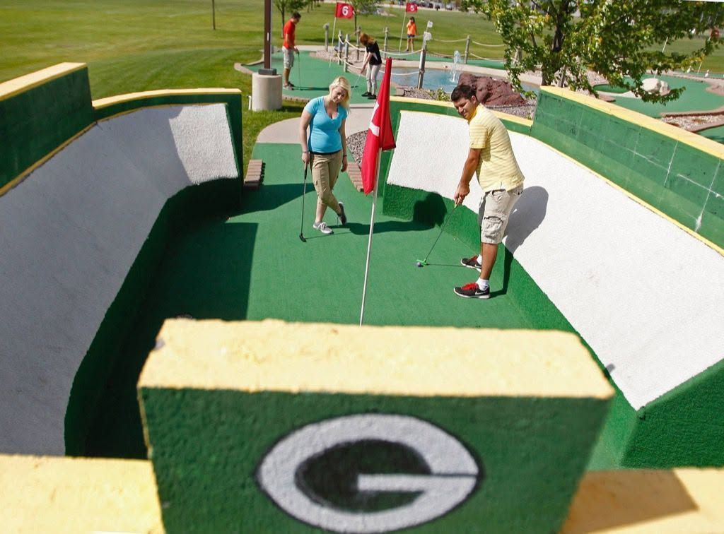 Two people putting on a green mini golf hole with a Green Bay Packers logo backdrop