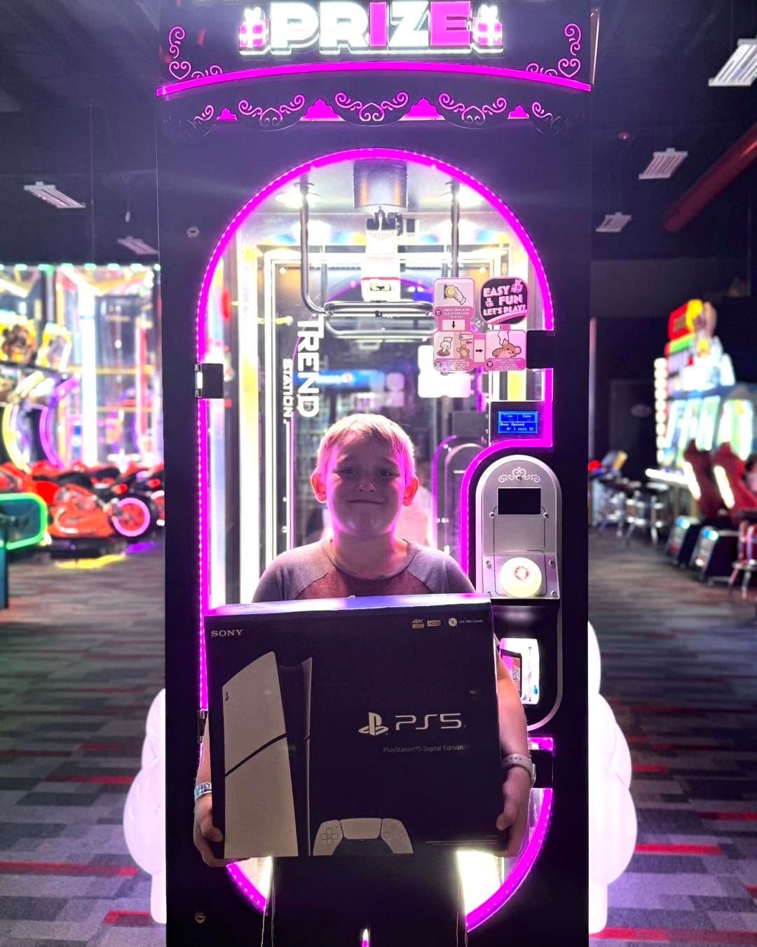 Person standing in a neon-lit arcade prize booth holding a PS5 box