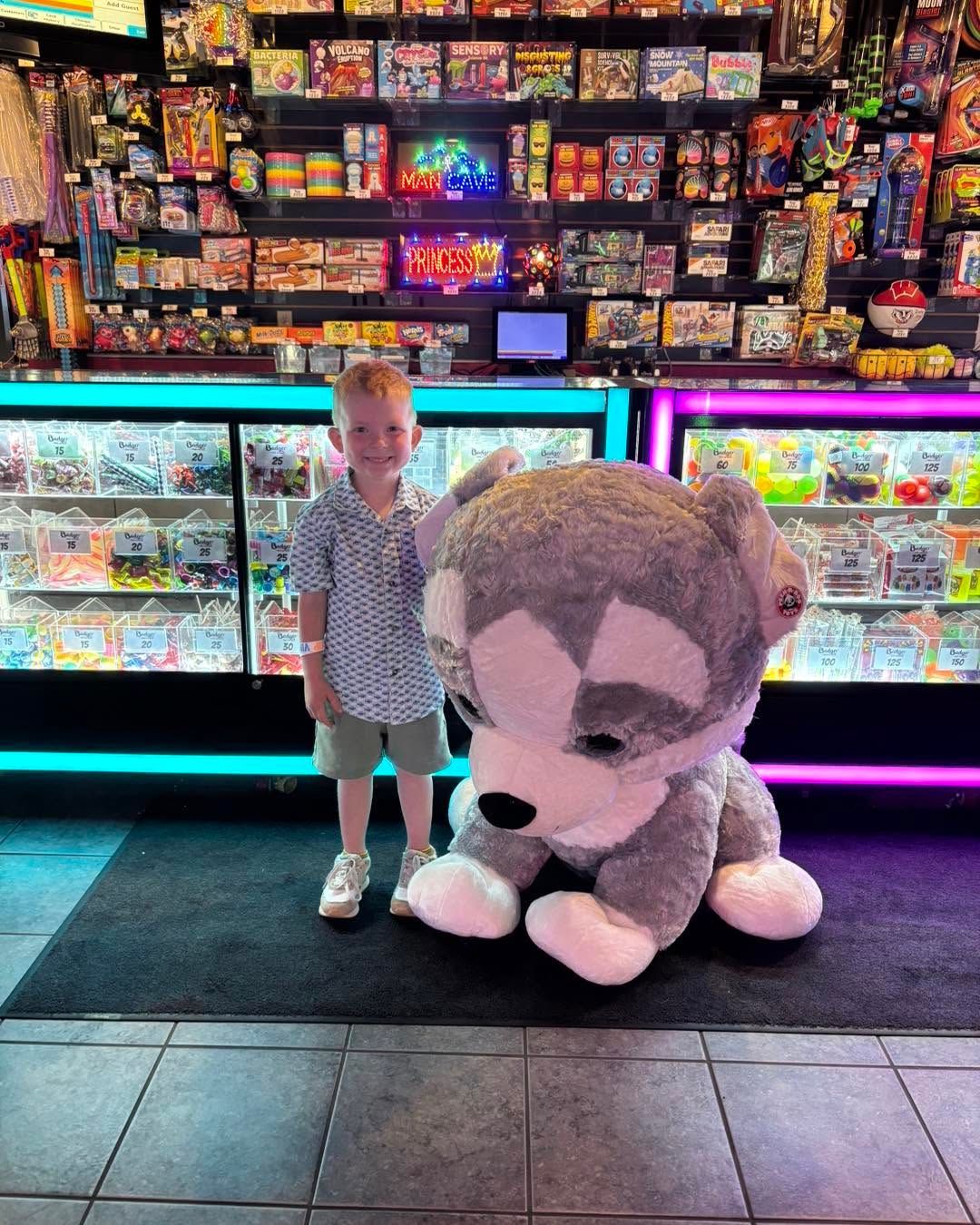 Child standing beside a giant plush toy in front of a brightly lit candy and snack display.
