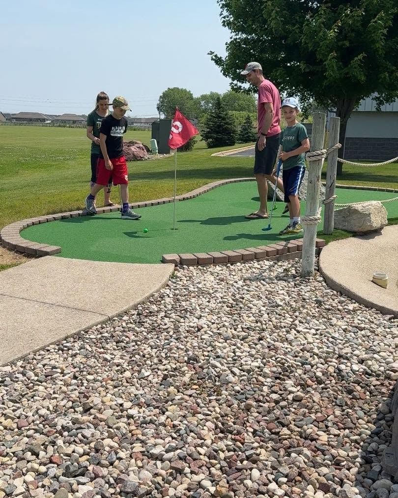 People playing mini golf on a green course with a red flag and rock borders on a sunny day