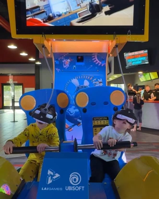 Two people play a blue Ubisoft arcade game with large controllers in a bright gaming venue.