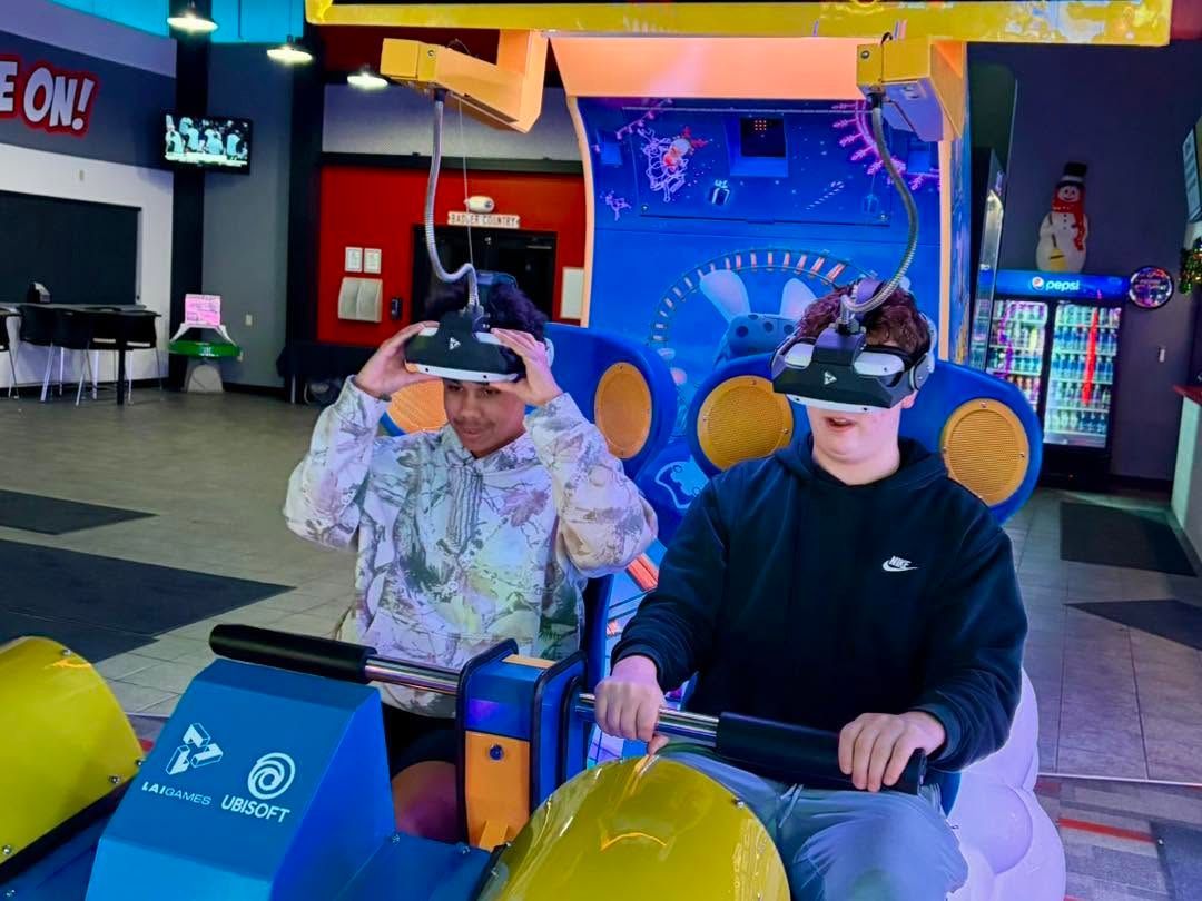 Two people wearing VR headsets sit at an arcade racing game, with bright colorful lights behind them.