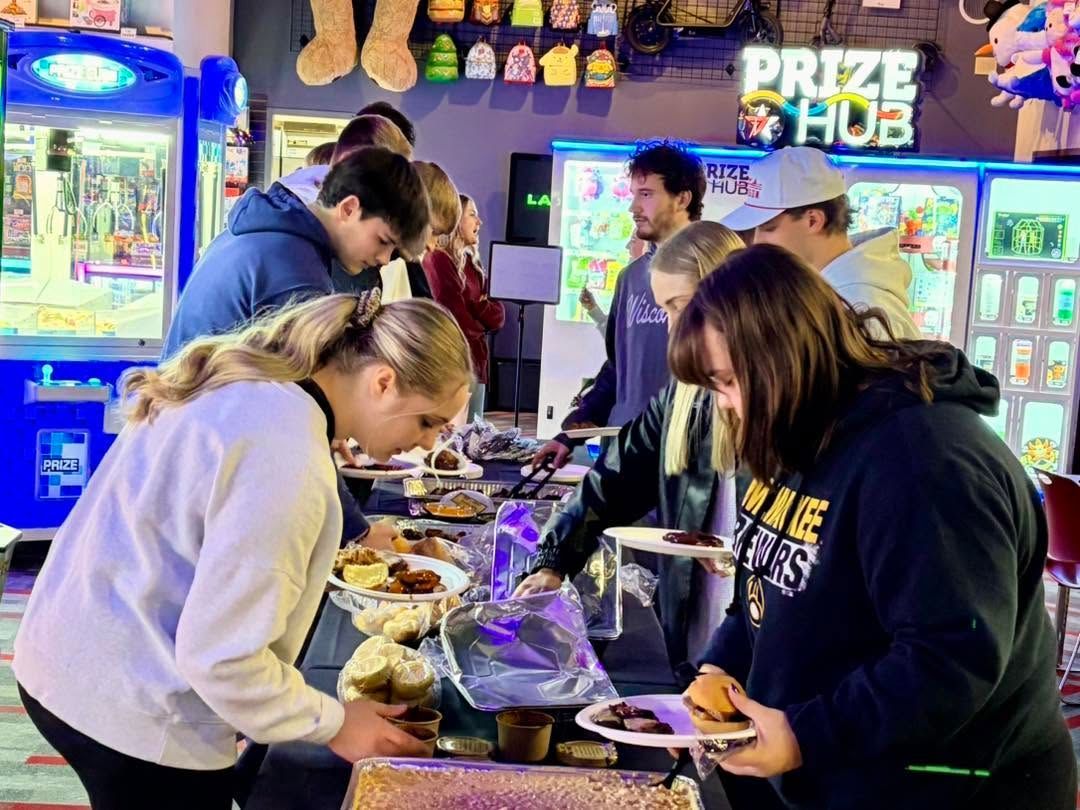 People serving food at a busy arcade buffet table under neon game lights