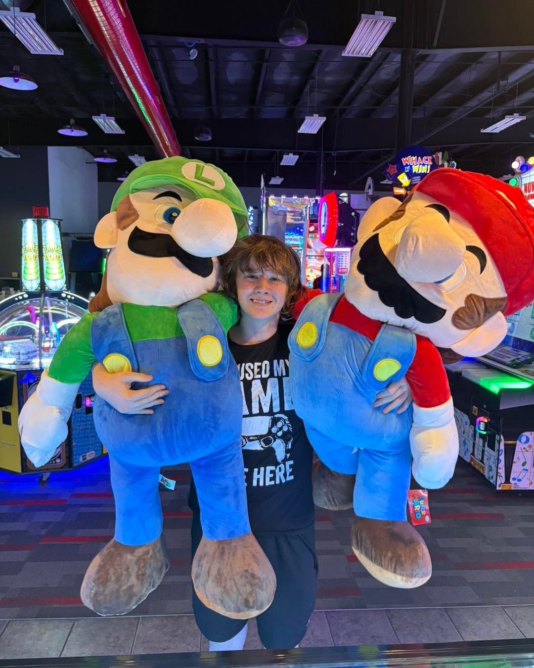 Person posing between two giant Luigi and Mario plush characters in a colorful arcade.