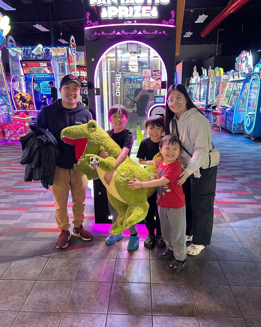 Family posing with a green dinosaur ride at a brightly lit arcade.