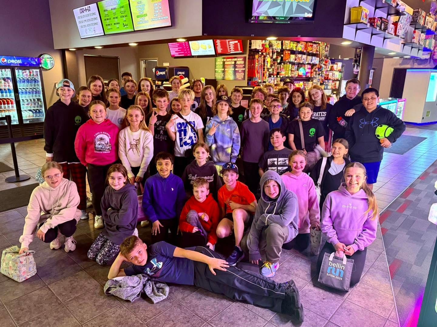 Large group posing inside a bowling alley, with colorful arcade screens and lanes in the background.