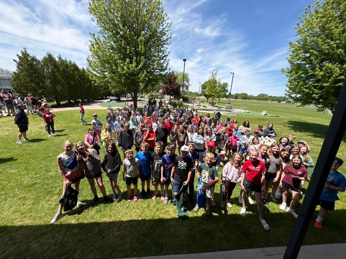 Large group gathered on a grassy field at an outdoor event under a bright blue sky.