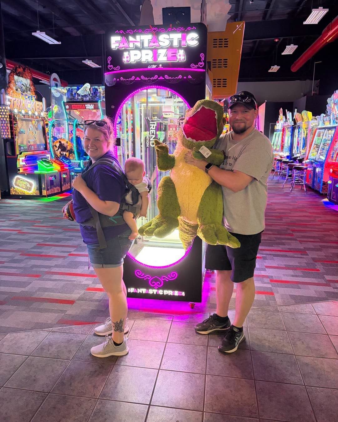 Three people posing with a dinosaur statue at a brightly lit arcade at night