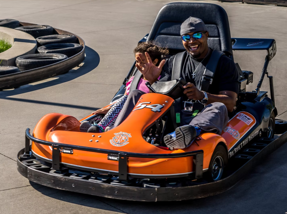 Two people ride an orange bumper car on a track, smiling and waving.