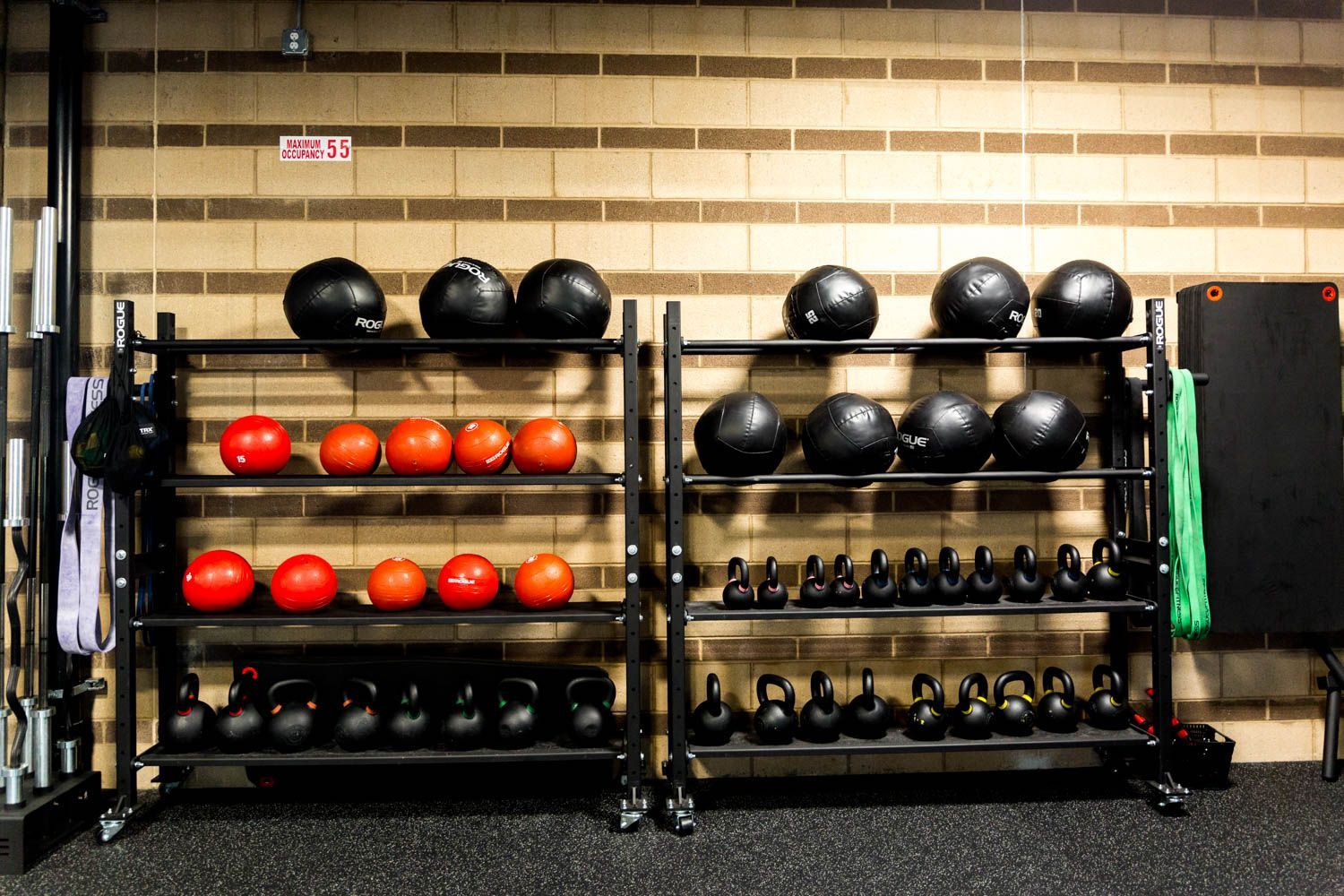 Gym equipment storage: black racks hold medicine balls, kettlebells, and dumbbells. Brick wall background.