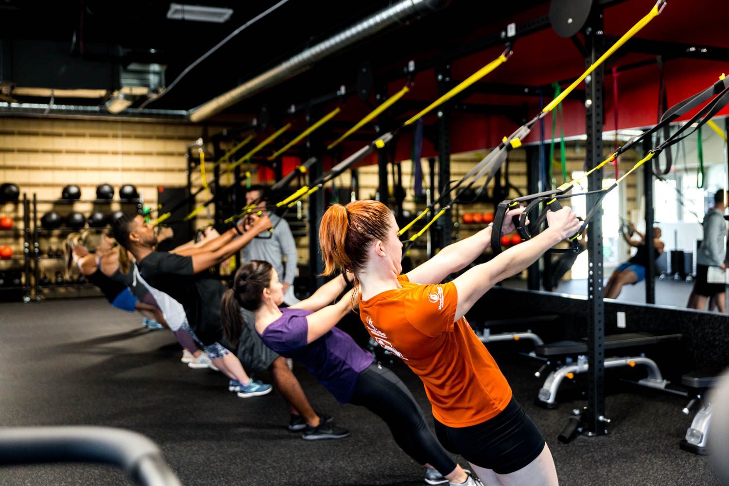 People exercising in a gym, using suspension trainers. Red and yellow straps hang from the ceiling.
